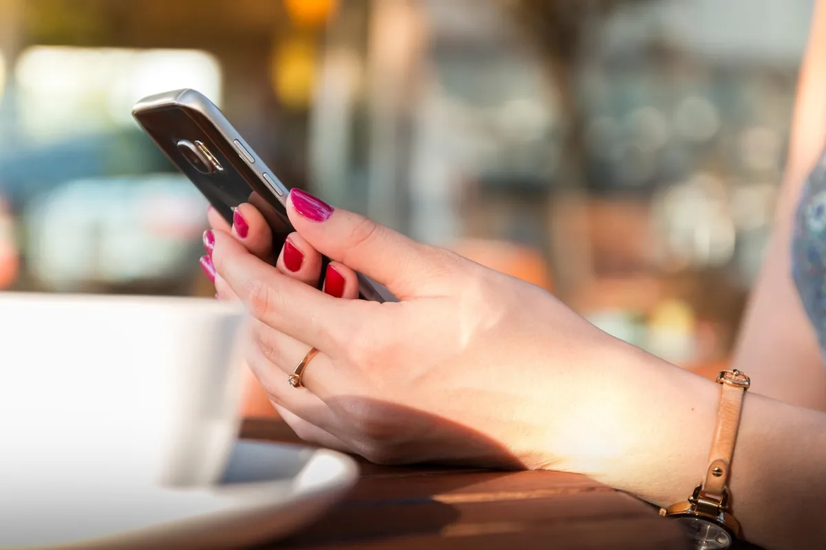 A hand holding a smartphone at a table, a cup beside it.
